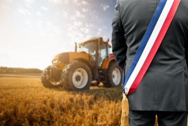 Maire avec son écharpe face à un champ de blé avec tracteur.